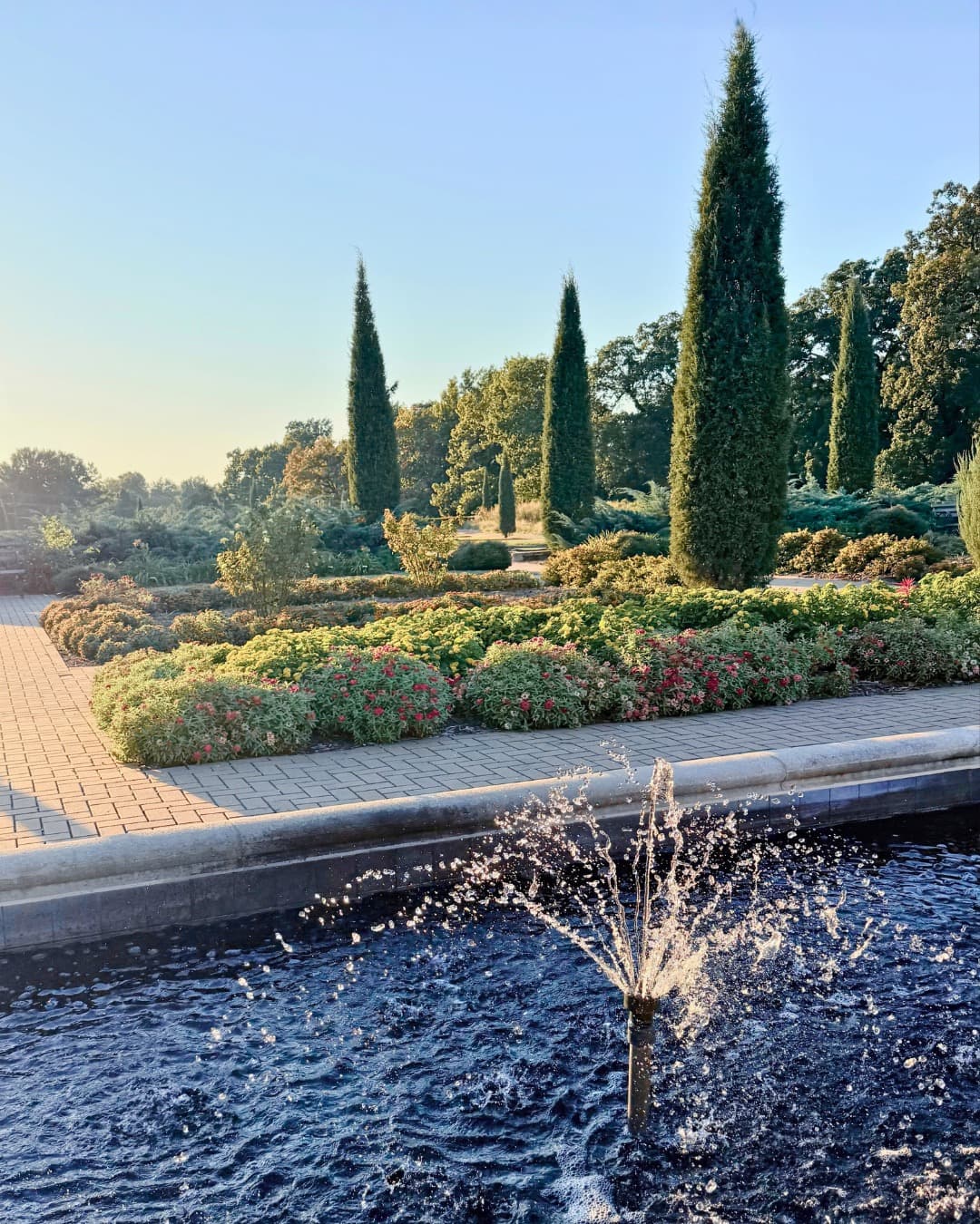 A fountain splashing in a long stone pool in the formal gardens of Woodward Park, with low flower beds, a brick walk and tall Italian cypresses beyond under a clear evening sky.
