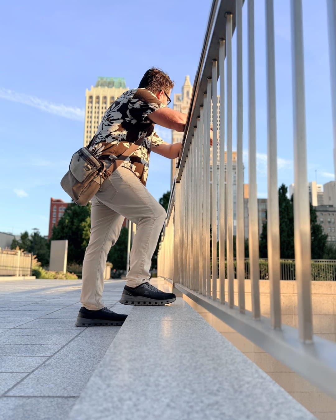 A man seen from behind, one foot up on the base of a downtown walkway railing, looking south toward the Philtower and the towers of Boston Avenue.